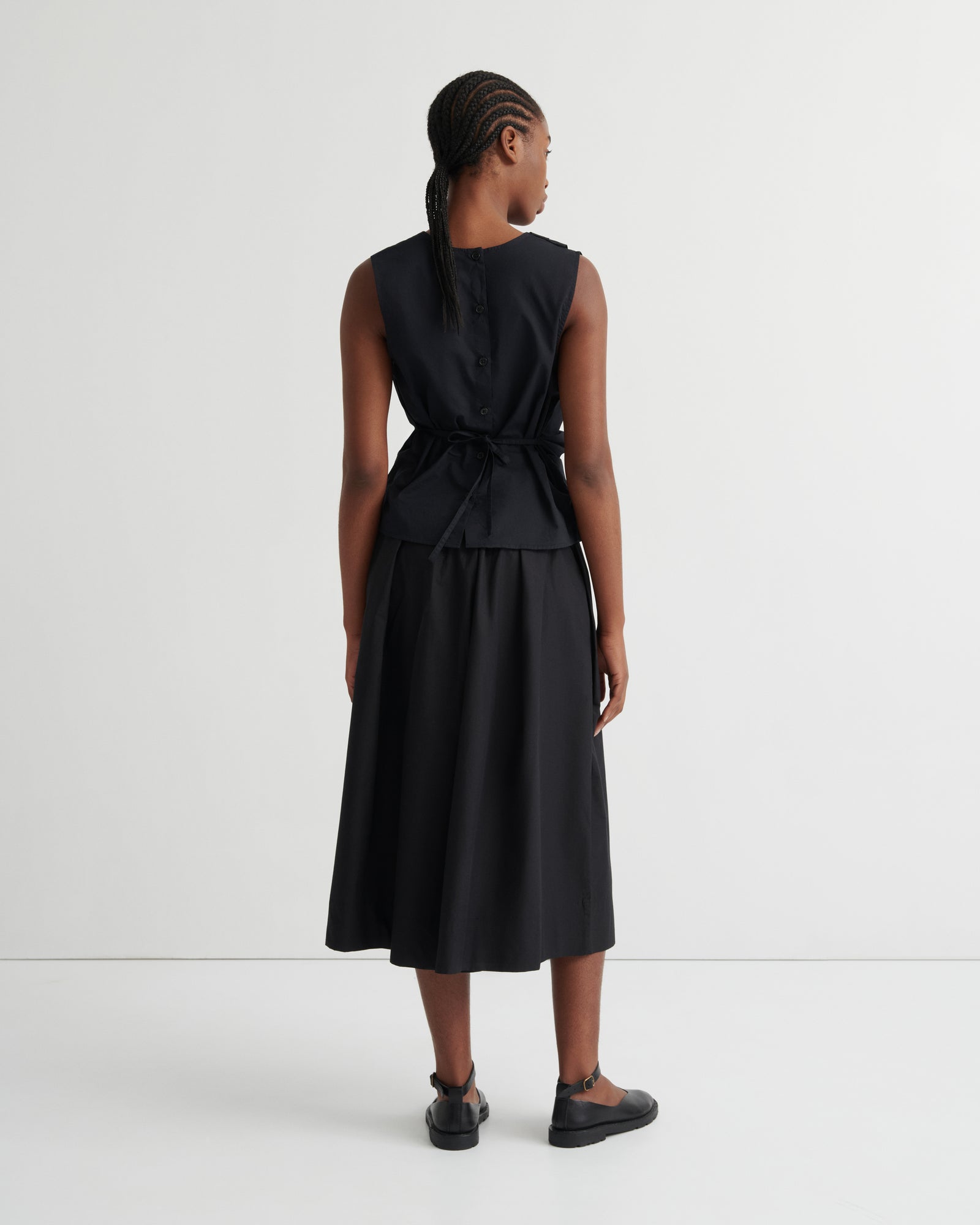 A model viewed from the back wearing a sleeveless black midi dress with a tie waist, paired with black flat shoes, against a plain white studio background.