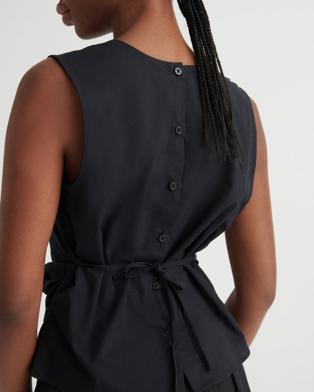 Close-up back view of a woman wearing a sleeveless black dress with a buttoned back and a self-tie waist sash, against a plain light background.