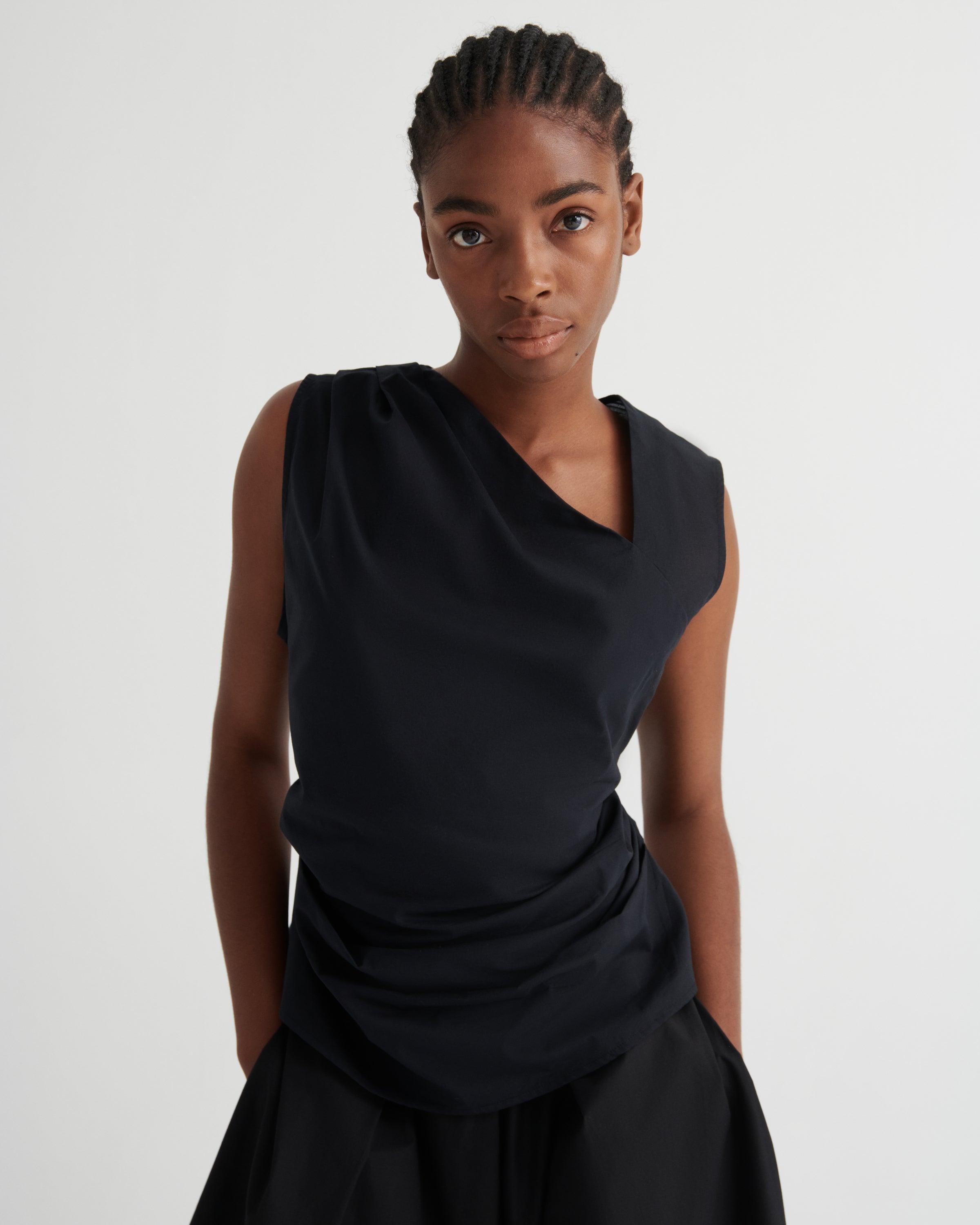 A model wearing a black draped sleeveless top and matching black skirt against a plain light gray studio background.