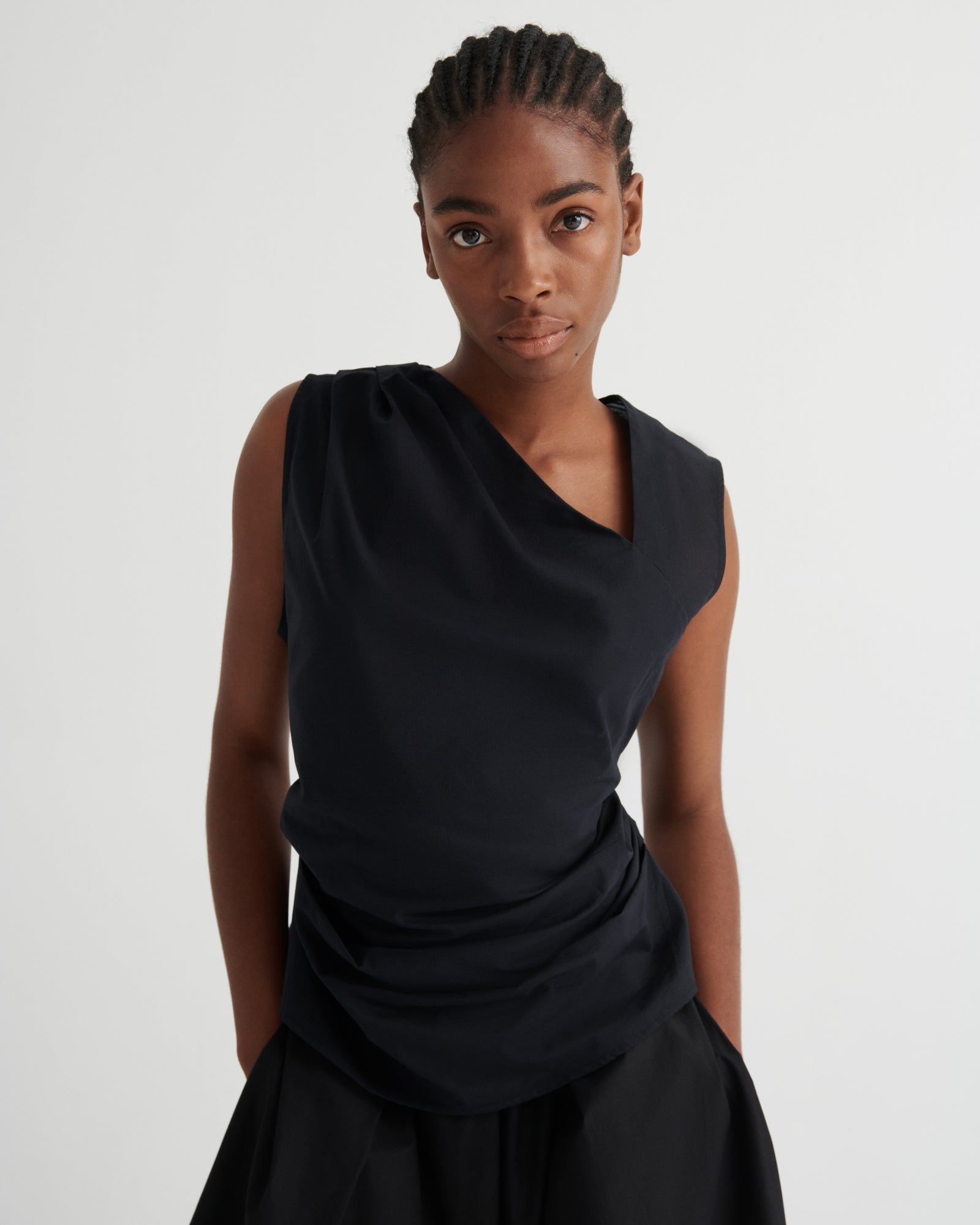 A model wearing a black draped sleeveless top and matching black skirt against a plain light gray studio background.