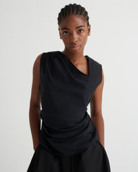 A model wearing a black draped sleeveless top and matching black skirt against a plain light gray studio background.