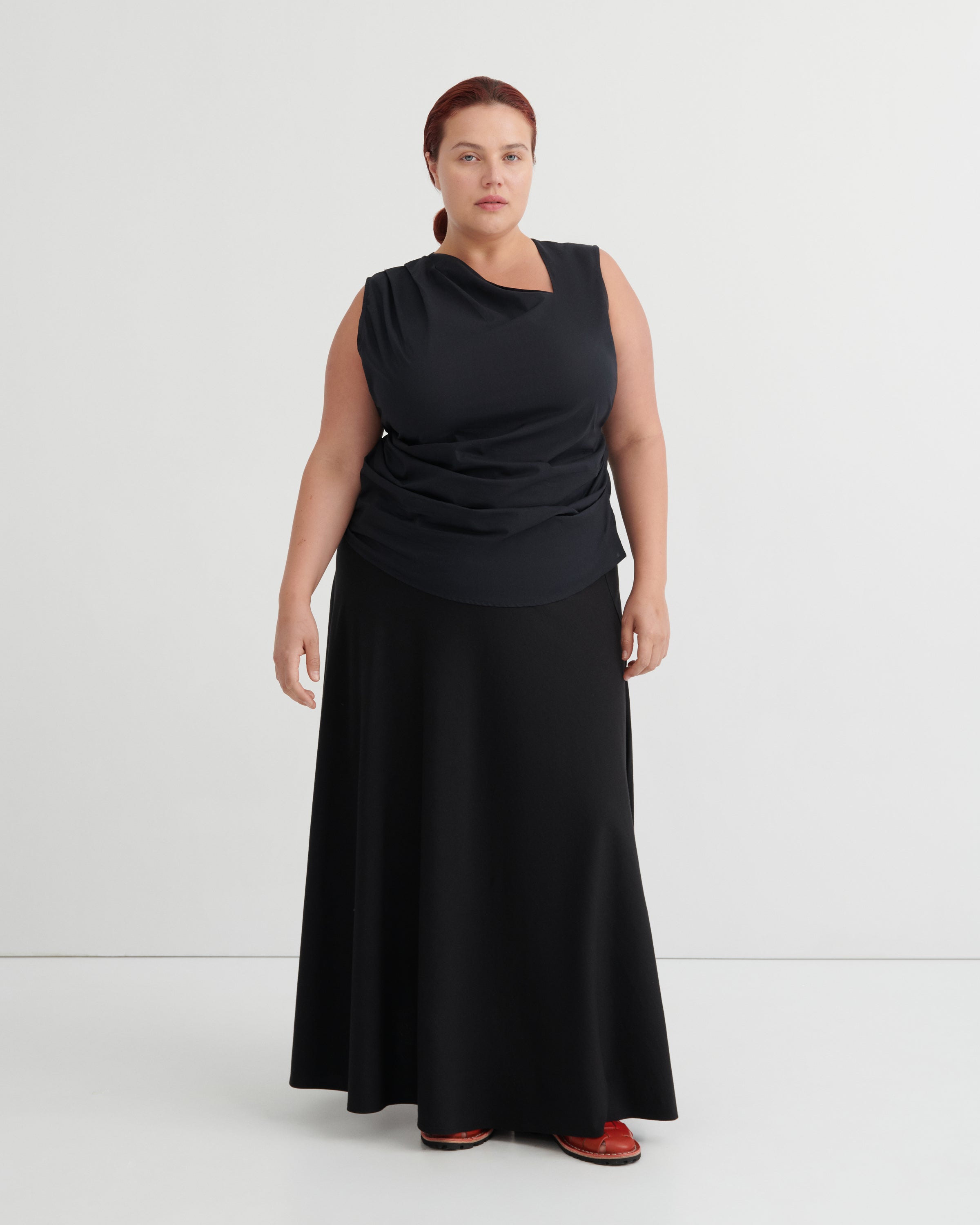 A woman modeling a sleeveless black draped top and matching long black skirt against a plain light gray studio background.