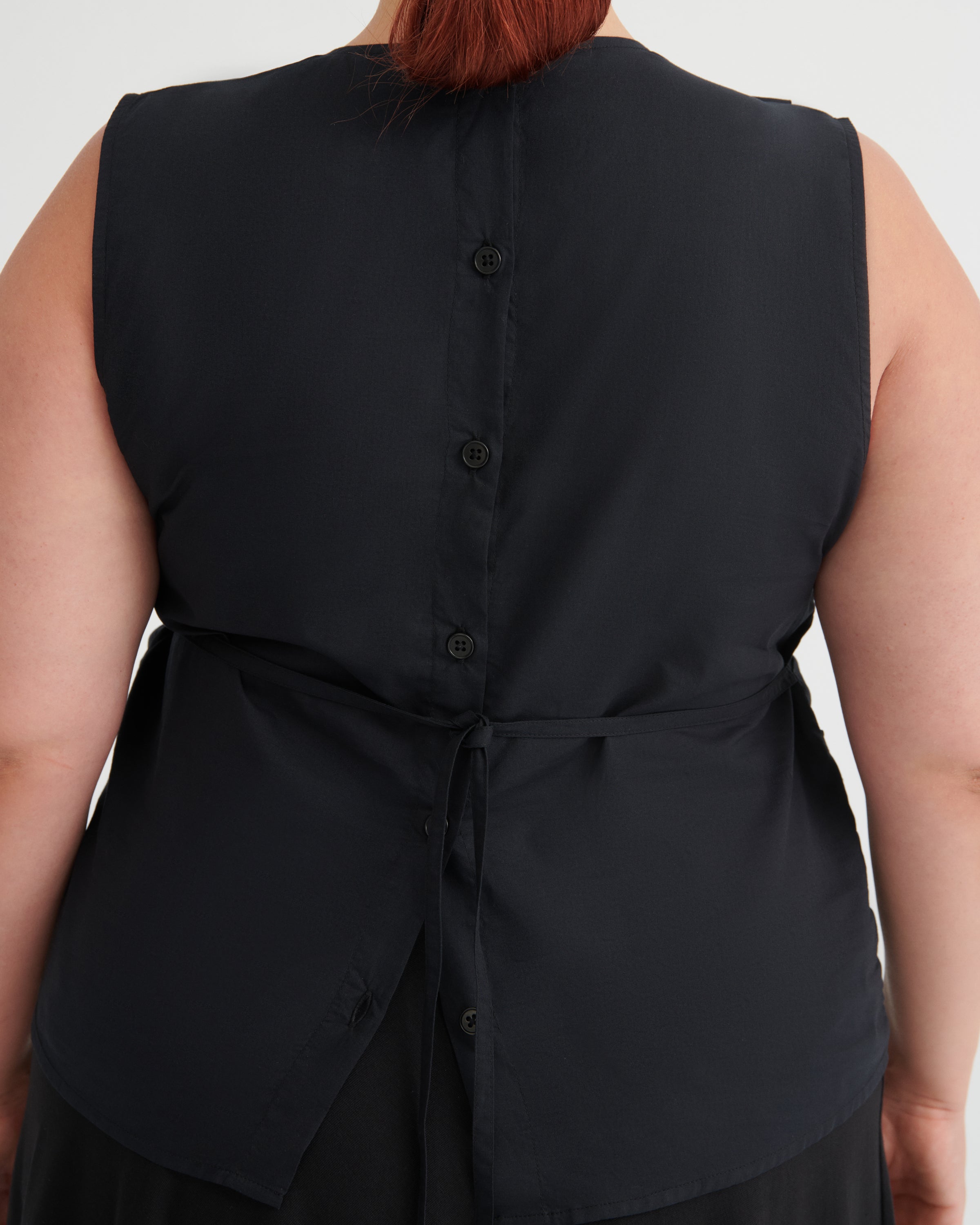 Close-up back view of a sleeveless black top with a buttoned back and tie detail, worn by a model against a light gray background.