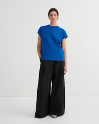 A model stands against a plain light gray studio background wearing a bright blue short-sleeve crew-neck T-shirt and wide-leg black trousers.