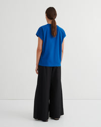 A woman is shown from behind wearing a bright blue short-sleeve top and black wide-leg pants against a plain white studio background.