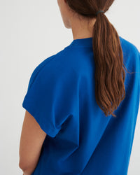 A woman shown from the back wearing a bright blue short-sleeve crewneck top, with her brown hair tied back in a ponytail against a plain light background.