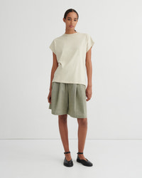 A model stands against a plain light gray studio background wearing a cream-colored short-sleeve t-shirt and olive green linen shorts, paired with black flat shoes.