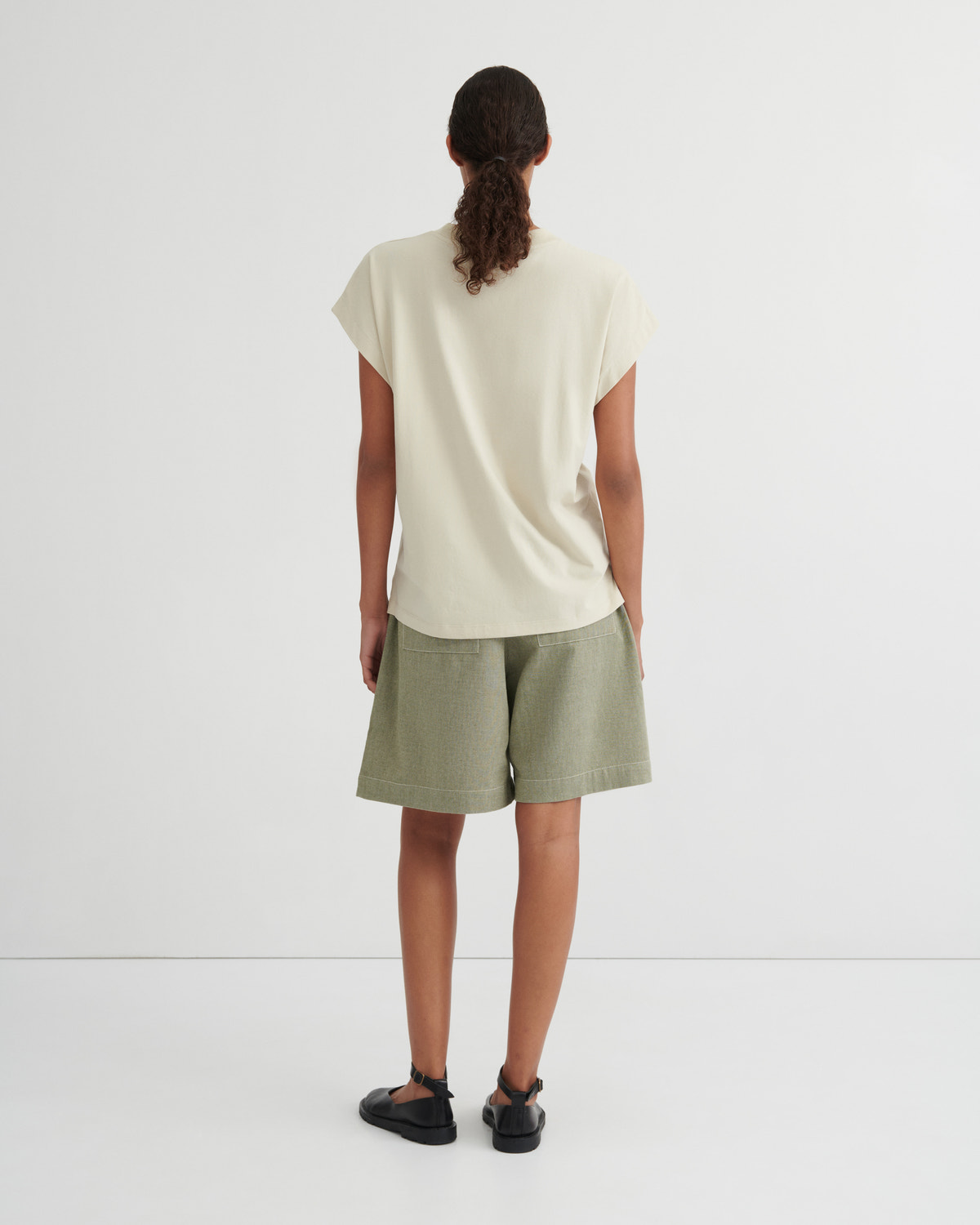 A model viewed from behind wearing a cream-colored short-sleeve t-shirt and sage green shorts against a plain white studio background.