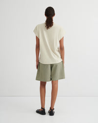 A model viewed from behind wearing a cream-colored short-sleeve t-shirt and sage green shorts against a plain white studio background.