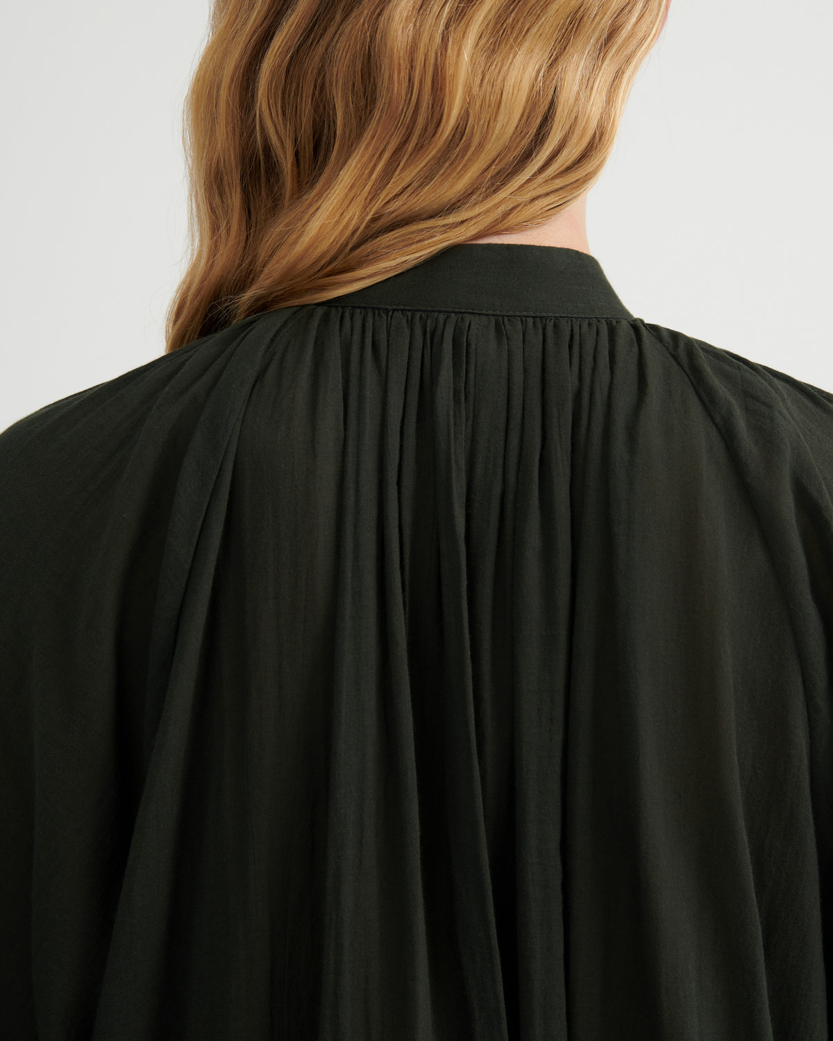 Close-up back view of a person wearing a dark olive green or black pleated blouse with a high gathered collar, shown against a plain light background.