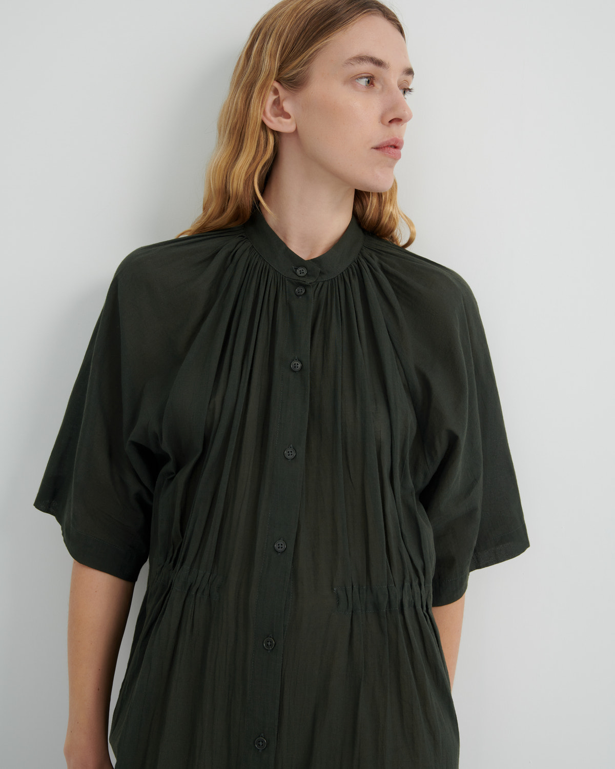 A woman wearing a dark olive green short-sleeve button-up dress with a high gathered collar and pleated details, photographed against a plain light gray wall.