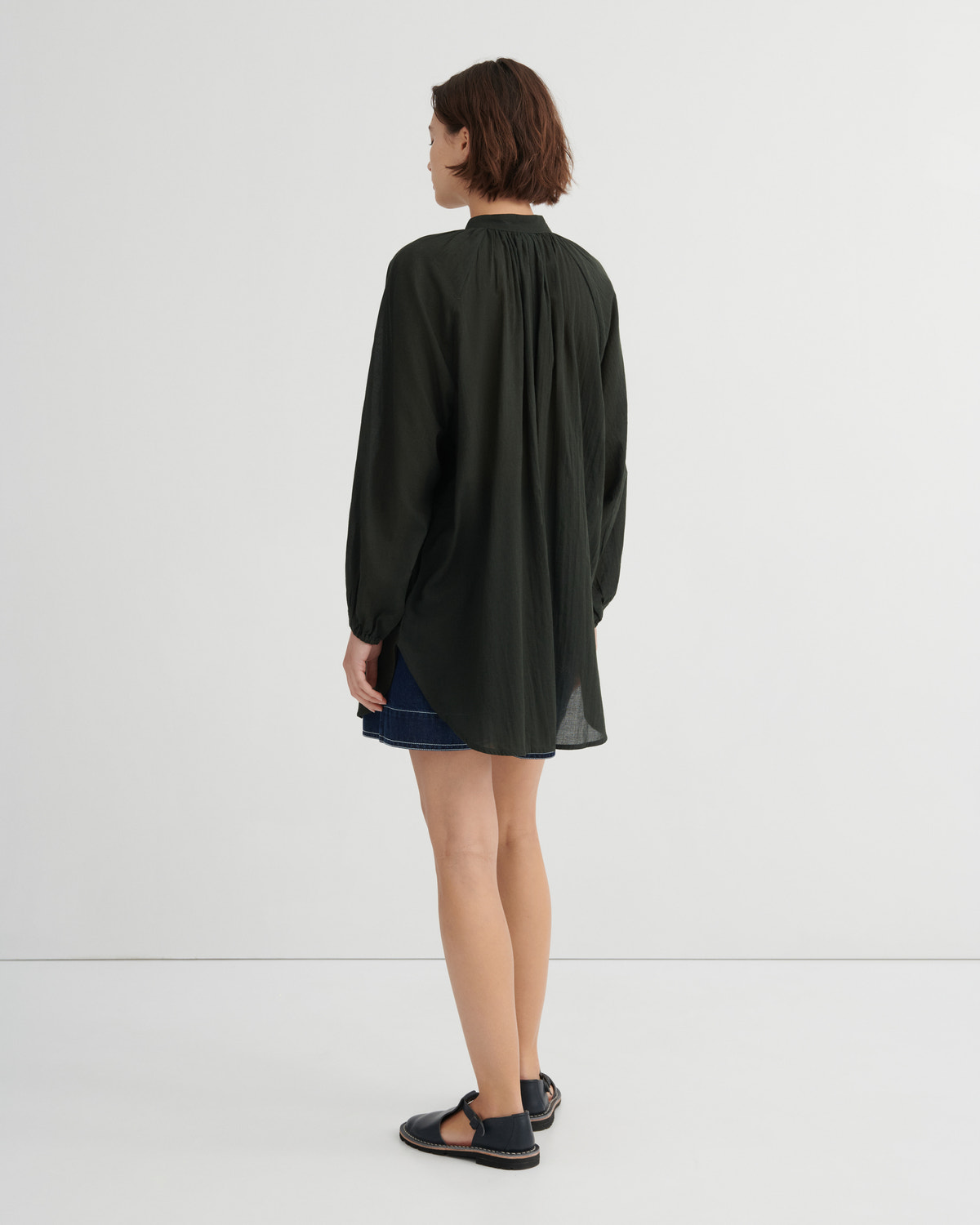 A woman shown from the back wearing a dark olive green long-sleeve blouse with a gathered neckline, paired with a dark denim skirt and black sandals, against a plain white studio background.