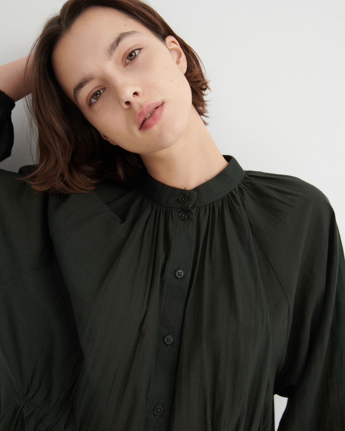 A close-up shot of a woman wearing a dark olive green long-sleeve blouse with a gathered high collar and button front, set against a plain light gray background.