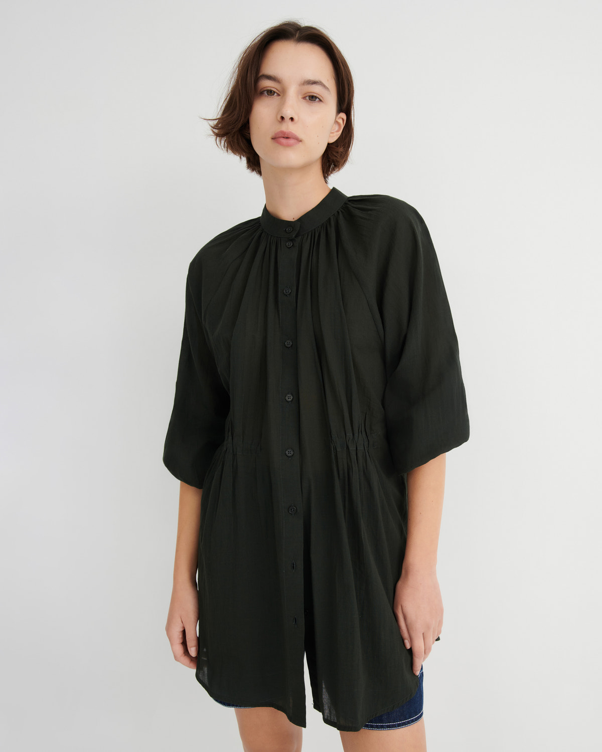 A woman modeling a black, oversized short-sleeve button-up shirt dress with a gathered high neck and side slits, paired with blue denim shorts, against a light gray studio background.