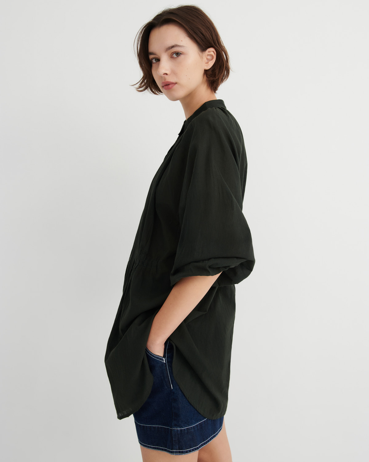 A woman wearing an oversized black short-sleeve button-up shirt with rolled cuffs and a dark denim skirt, photographed against a plain light gray studio background.