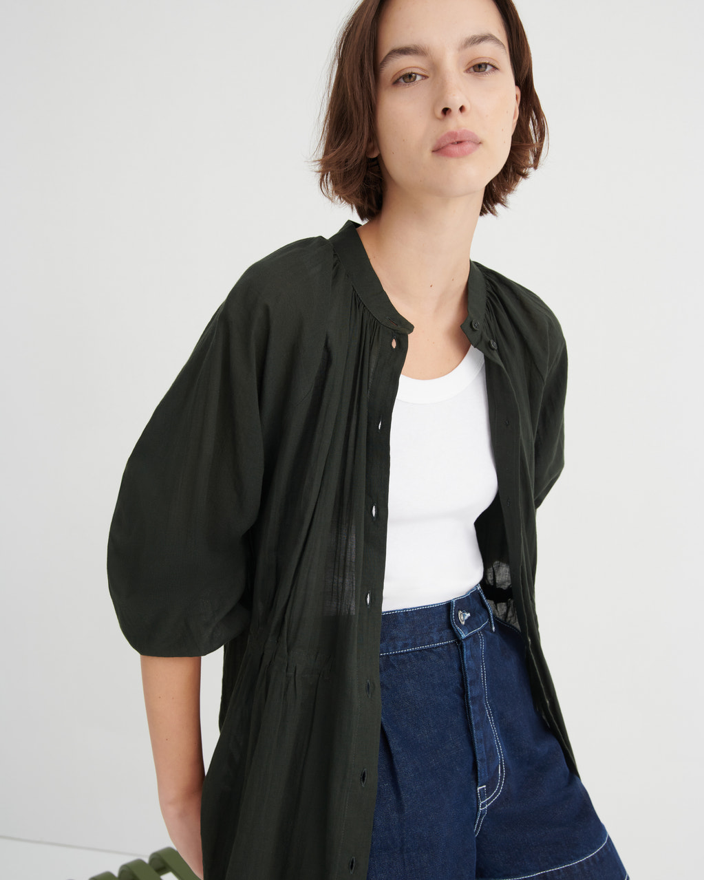 A woman wearing a dark olive green button-up shirt jacket with voluminous puff sleeves over a white tank top and blue jeans, photographed against a clean white studio background.