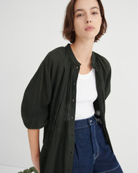 A woman wearing a dark olive green button-up shirt jacket with voluminous puff sleeves over a white tank top and blue jeans, photographed against a clean white studio background.
