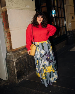 A woman stands on a city sidewalk at night wearing a bright red long-sleeve top and a long gray skirt with a yellow and purple floral print. She is holding a small woven straw bag.