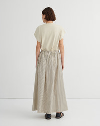 A woman viewed from behind wearing a cream short-sleeve top and a high-waisted, wide-leg pleated skirt with thin vertical stripes in beige and white. She is standing against a clean, off-white studio background.