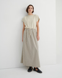 A model stands against a white wall wearing a cream-colored short-sleeve top and a matching beige and white vertically striped pleated maxi skirt with a drawstring waist, paired with black slip-on shoes.