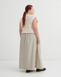 A woman with red hair in a ponytail is shown from the back wearing a cream sleeveless knit vest and beige-and-white vertically striped wide-leg trousers, standing against a plain white studio background.