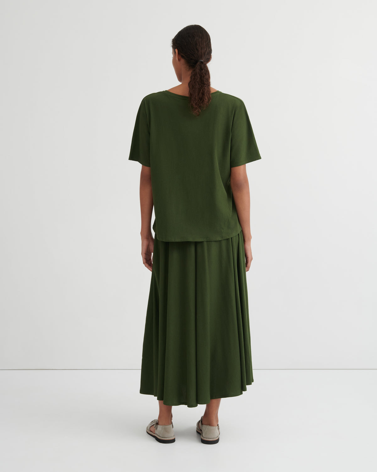 A woman viewed from the back wearing an olive green short-sleeve top and matching pleated midi skirt, paired with beige flat sandals, against a plain light gray studio background.