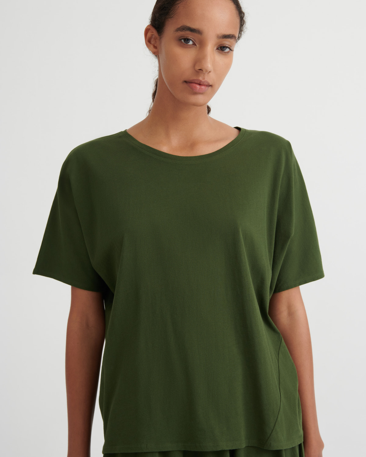A woman wearing a loose, short-sleeve crewneck t-shirt in olive green, photographed against a plain light gray studio background.
