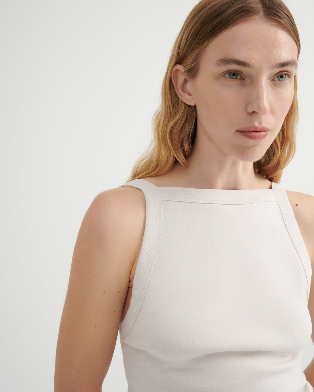 A close-up of a woman wearing a sleeveless, boat-neck top in a warm off-white or cream color.