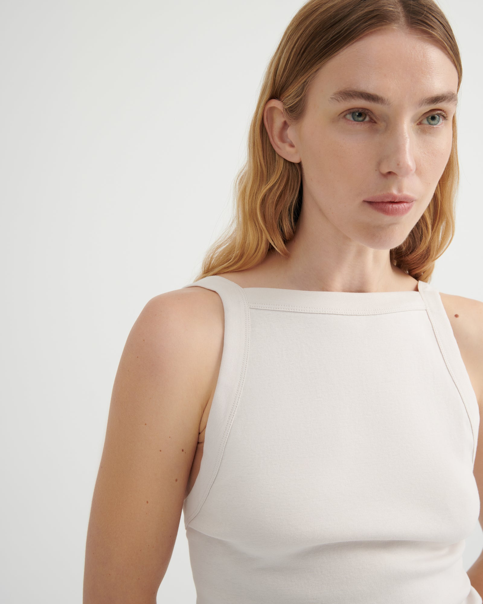 A close-up of a woman wearing a sleeveless, boat-neck top in a warm off-white or cream color.