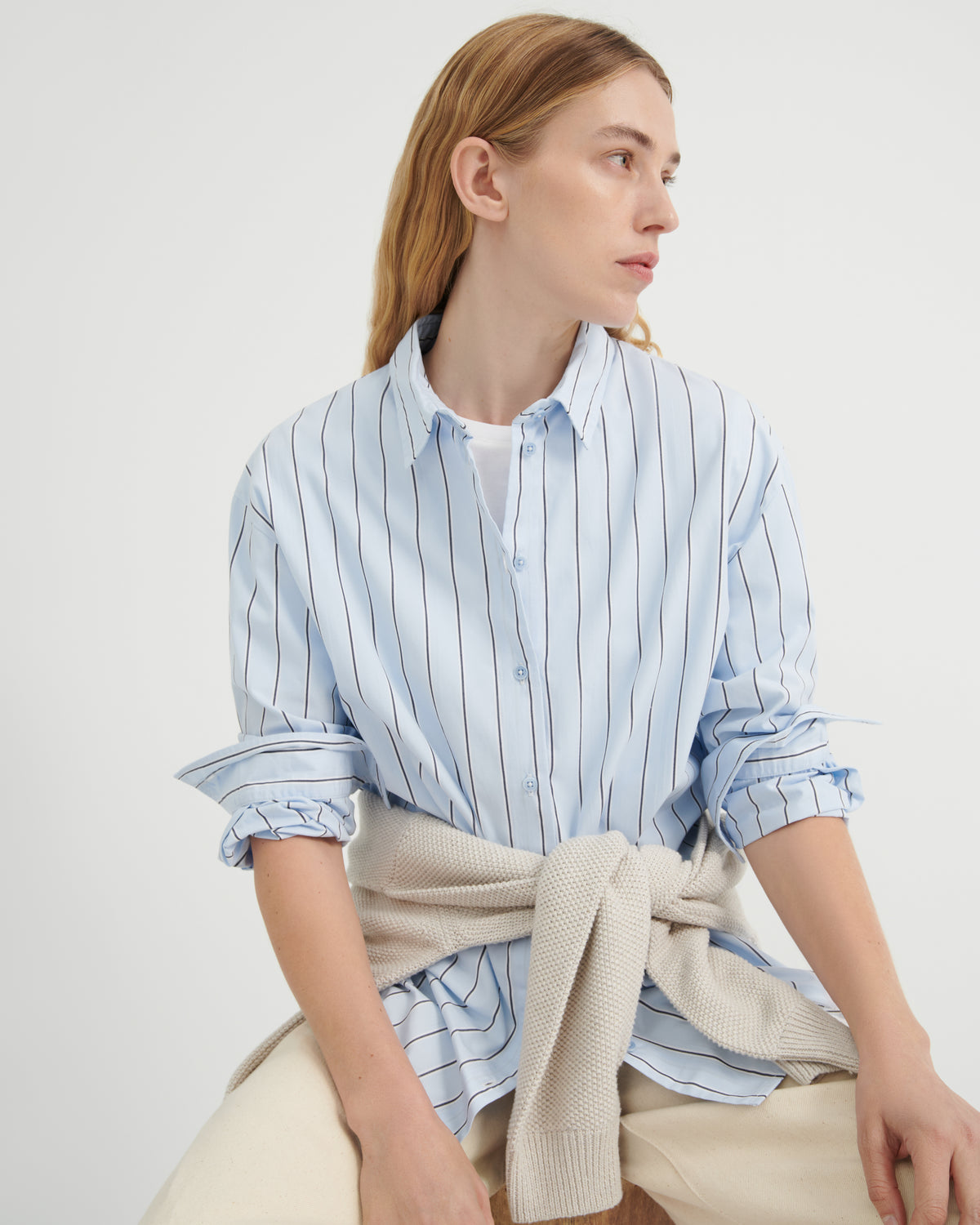 A woman wearing a light blue and white vertically striped button-down shirt with rolled-up sleeves, tied with a beige knit sweater.