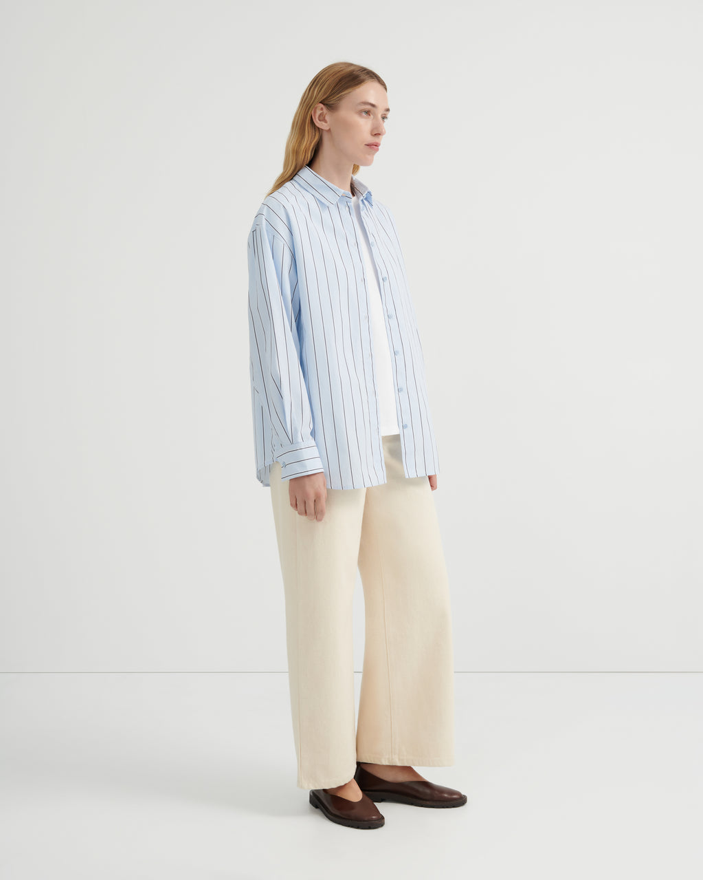 A woman stands in profile wearing a light blue and white vertically striped button-up shirt, cream wide-leg trousers, and dark brown loafers.