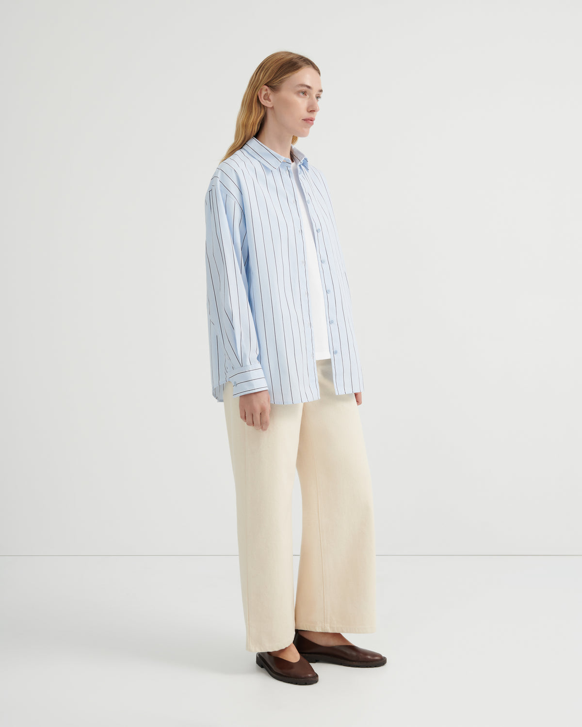 A woman stands in profile wearing a light blue and white vertically striped button-up shirt, cream wide-leg trousers, and dark brown loafers.