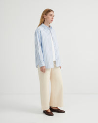 A woman stands in profile wearing a light blue and white vertically striped button-up shirt, cream wide-leg trousers, and dark brown loafers.