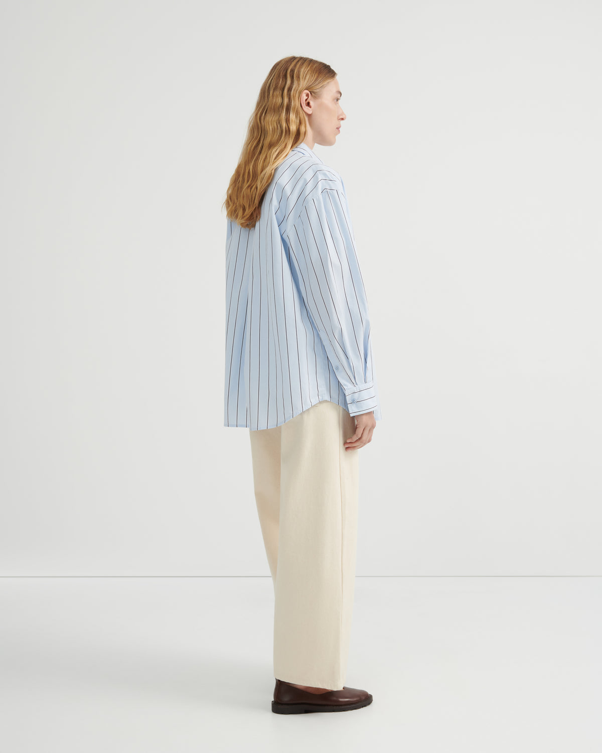 A woman shown from the side profile wearing a light blue and white vertically striped button-up shirt, cream wide-leg trousers, and dark brown leather loafers.