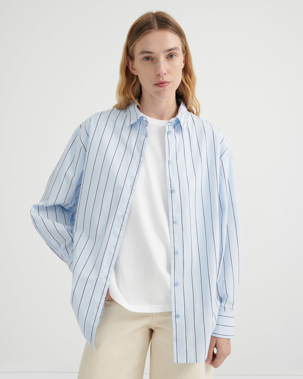 A woman wearing a light blue and white vertically striped button-up shirt over a white t-shirt and beige trousers.