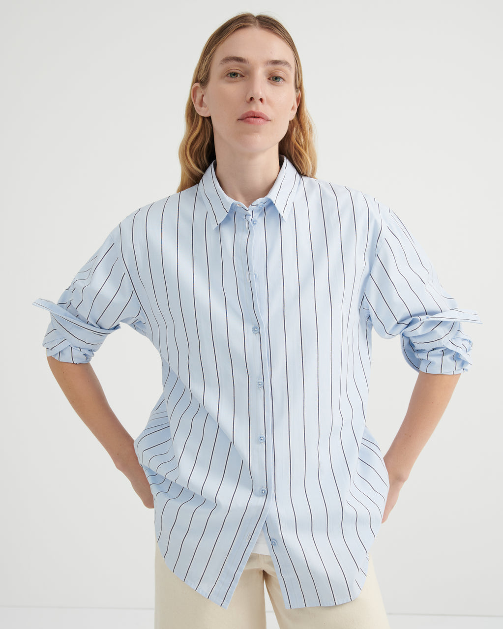 A woman modeling a light blue and white vertically striped button-down shirt with rolled-up sleeves, paired with cream pants.