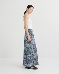 A woman stands in profile wearing a sleeveless white top and a long, wide skirt with an abstract blue, green, and purple marbled print. She is photographed against a clean light gray studio background.