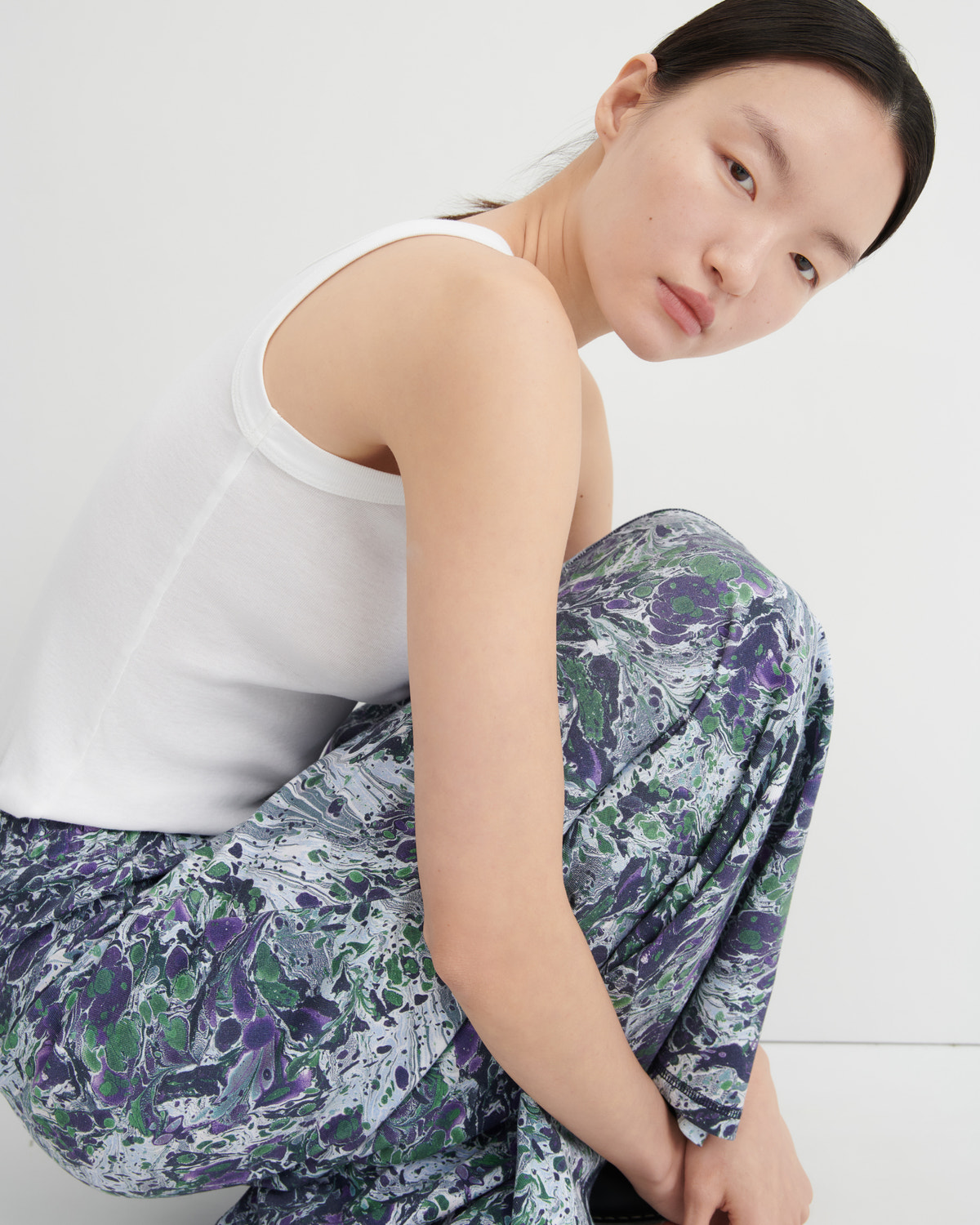 A model wearing a white ribbed tank top and loose-fit pants with a purple, green, and gray marble-like print, crouching against a plain light gray background.