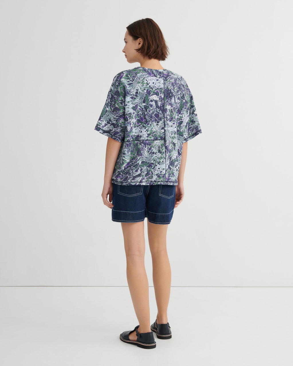 A woman shown from the back wearing a short-sleeve button-up shirt with a blue, green, and white marbled pattern, paired with dark denim shorts and black sandals, against a plain light gray studio background.