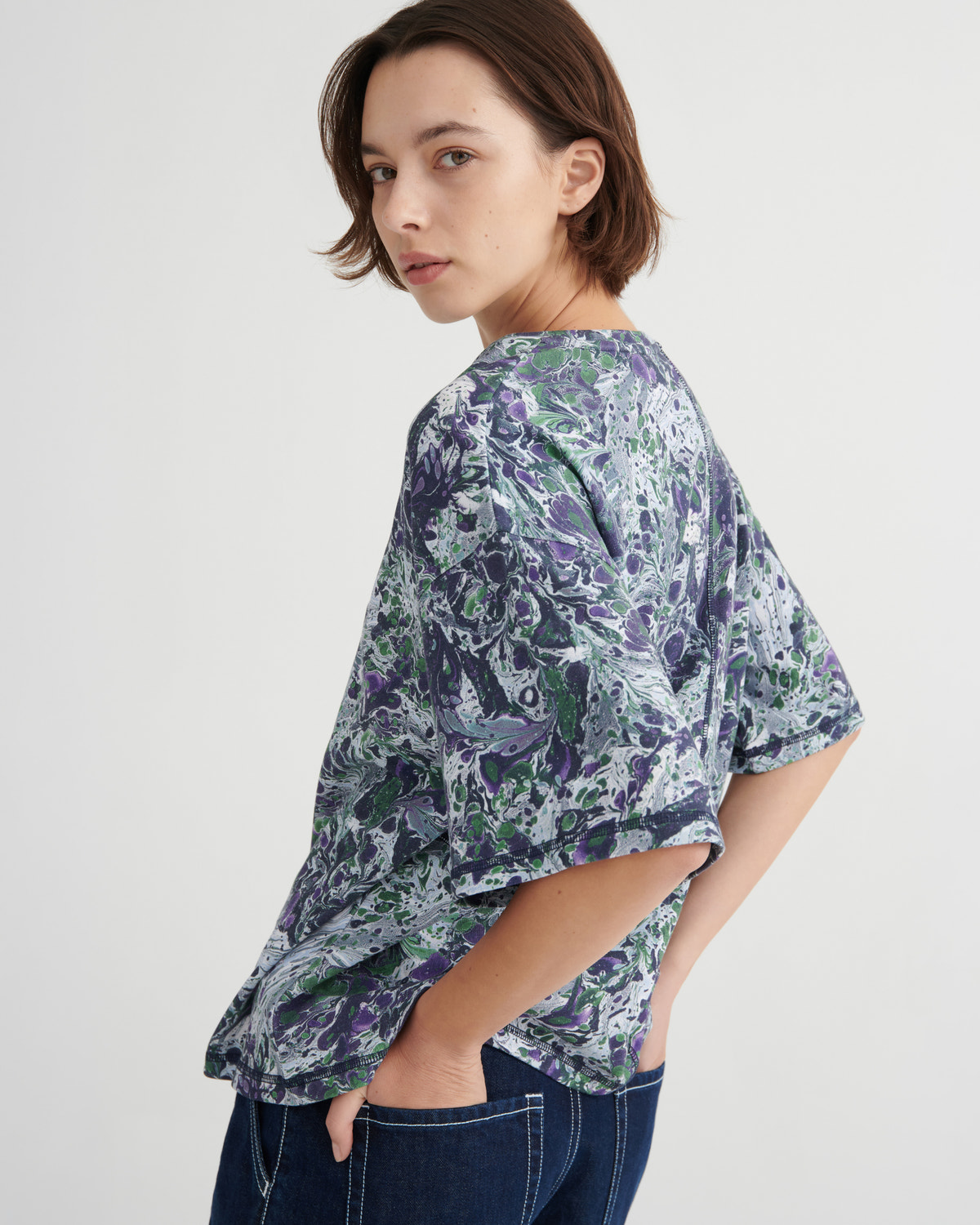 A woman wearing a loose short-sleeve top with a blue, purple, and green abstract marbled print, paired with dark blue jeans, photographed against a light gray studio background.