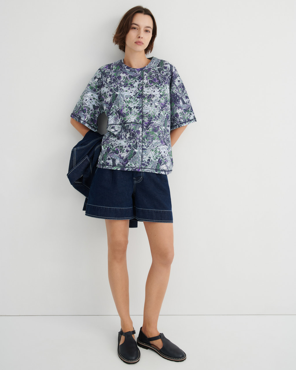 A model stands against a plain light gray studio wall wearing a short-sleeve button-front shirt with a blue, green, and white marbled print, dark denim shorts, and black slip-on shoes. A dark blue jacket is draped over one shoulder.