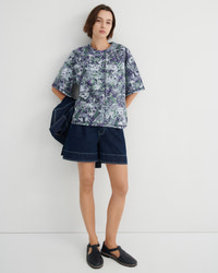 A model stands against a plain light gray studio wall wearing a short-sleeve button-front shirt with a blue, green, and white marbled print, dark denim shorts, and black slip-on shoes. A dark blue jacket is draped over one shoulder.