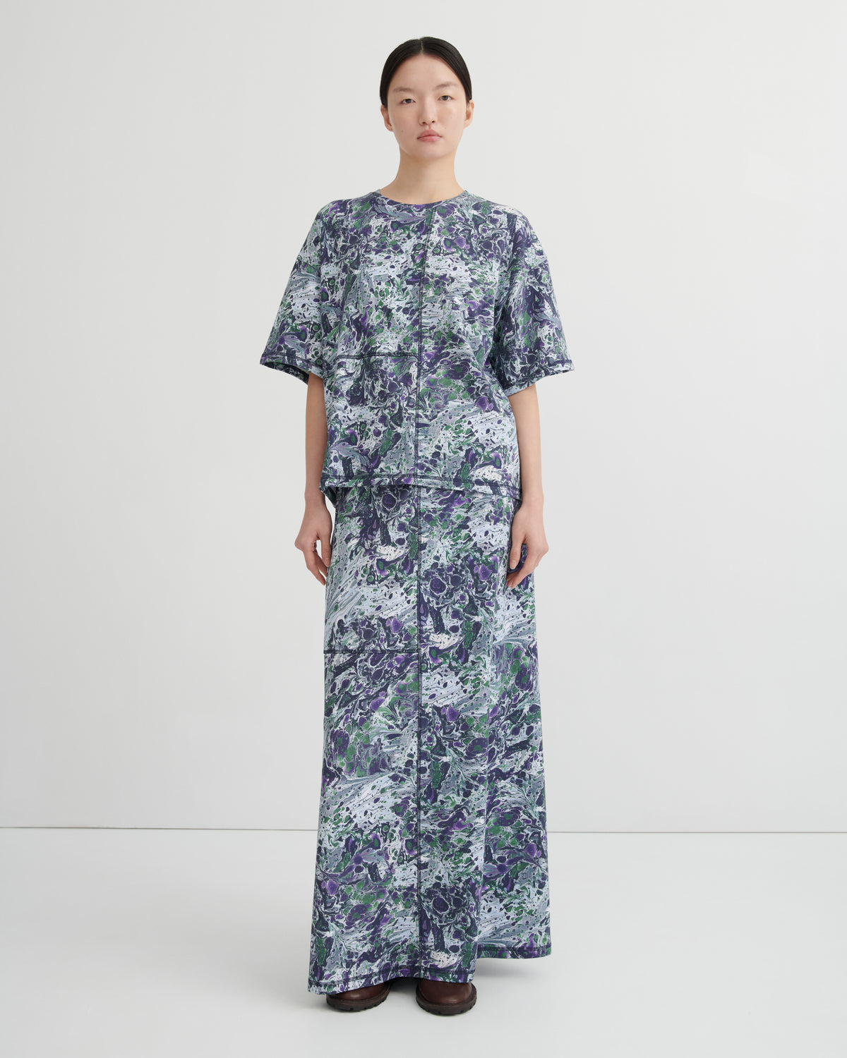 A model stands against a plain light gray studio background wearing a tee and maxi skirt with a blue, purple, and green abstract marbled pattern.