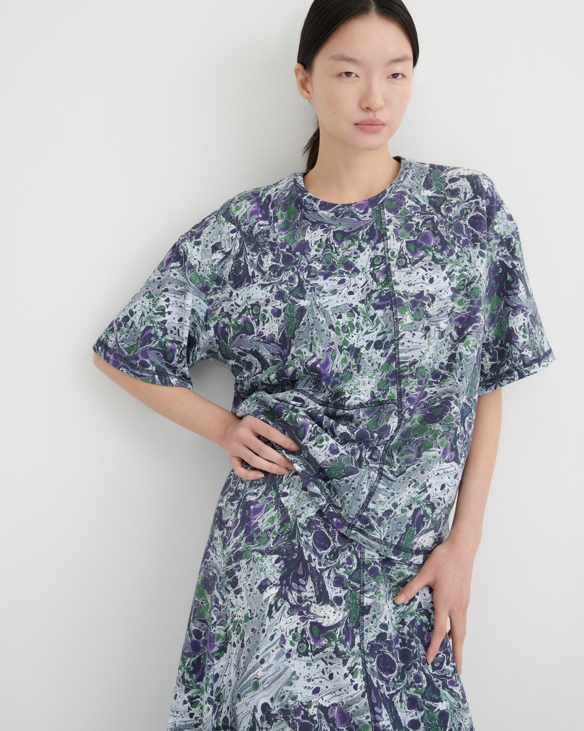 A model wearing a short-sleeve tee and maxi skirt with a purple, green, and white abstract marbled pattern, standing against a plain light gray wall.