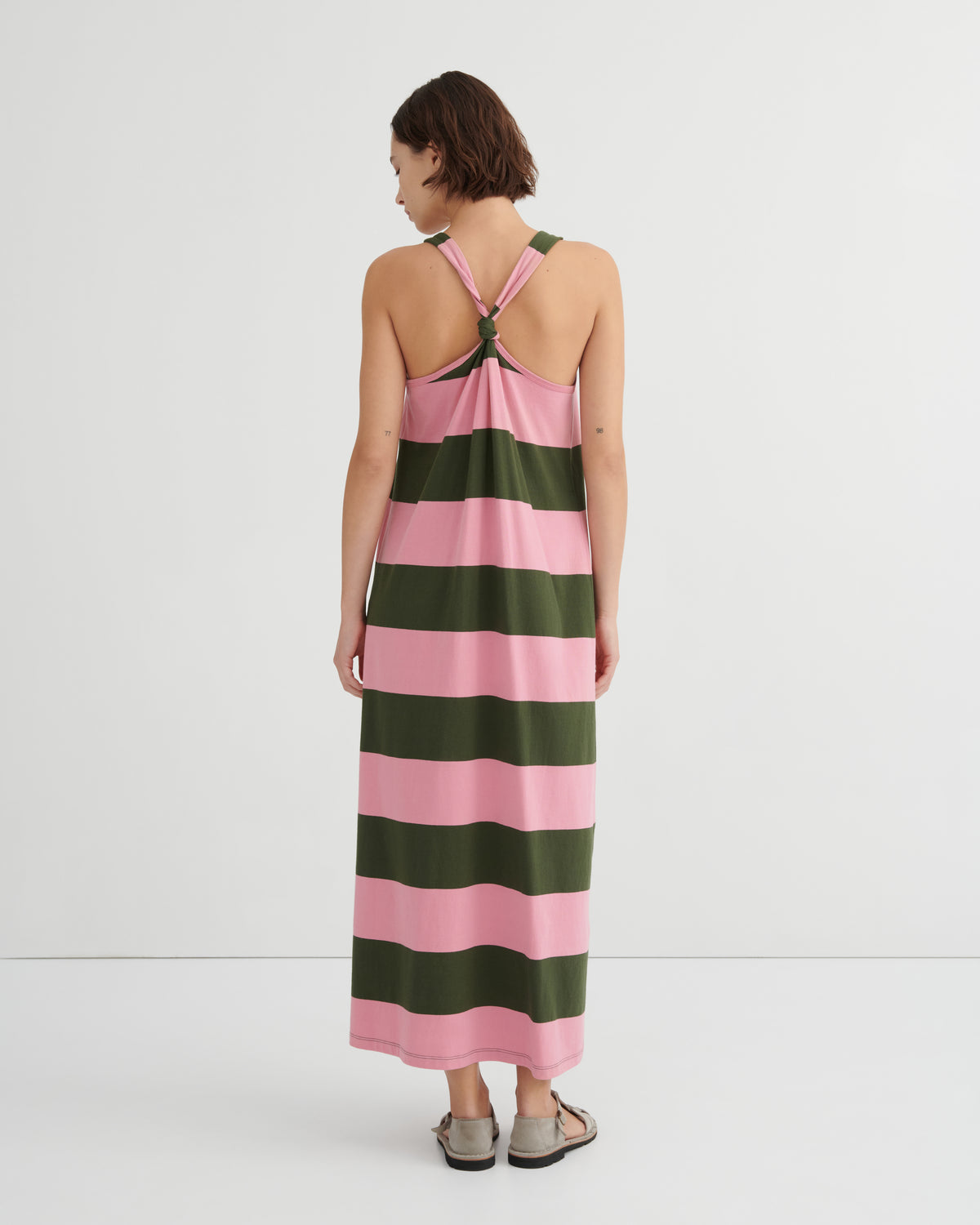 A woman viewed from the back wearing a sleeveless maxi dress with wide horizontal pink and dark green stripes and a crossover halter-style back, against a clean white studio background.