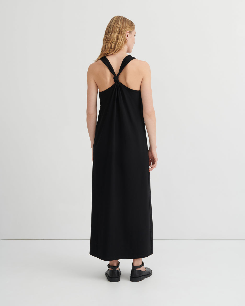 A woman viewed from the back wearing a sleeveless black maxi dress with a crossover back design, paired with black flat shoes, against a plain light gray studio background.