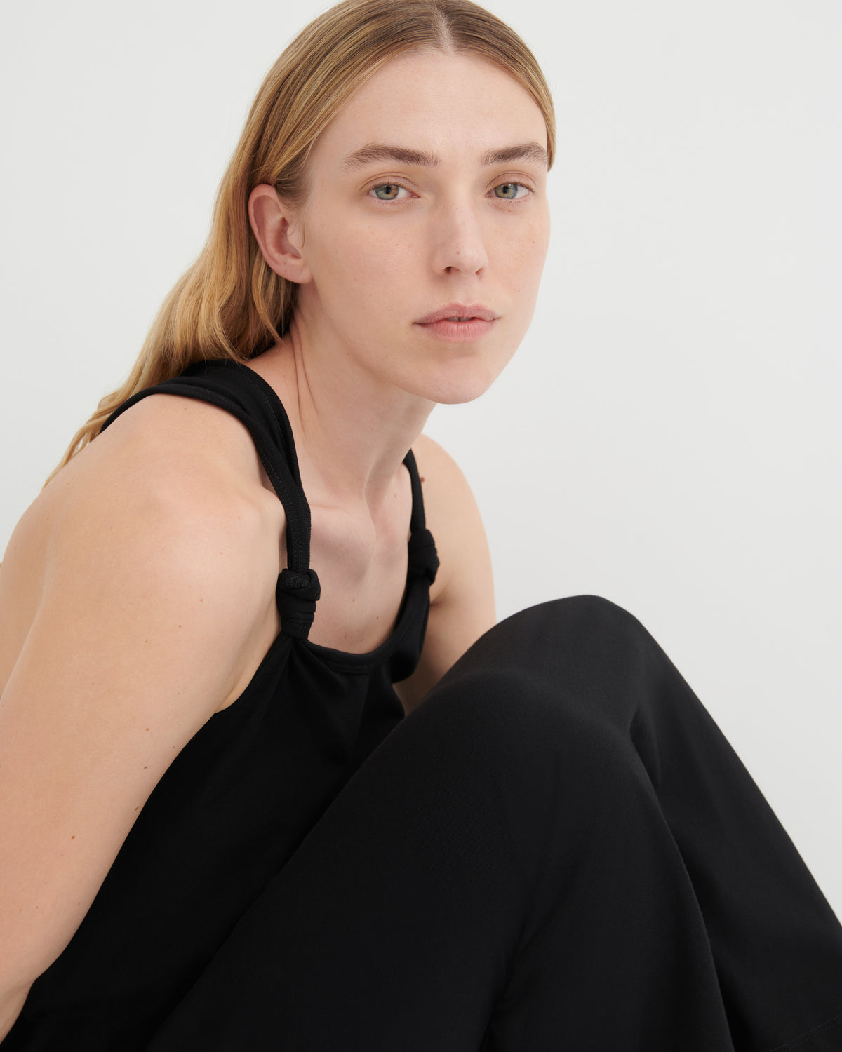 A woman wearing a black sleeveless top with knotted shoulder straps and black pants, photographed against a clean light gray studio background.