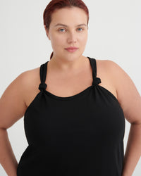 A plus-size model with red hair tied back, wearing a black halter-neck tank top with knotted shoulder details, photographed against a light gray studio background.
