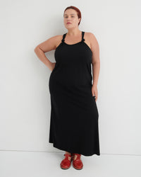 A woman modeling a black sleeveless maxi dress with thin shoulder straps and red shoes, standing against a plain white studio background.