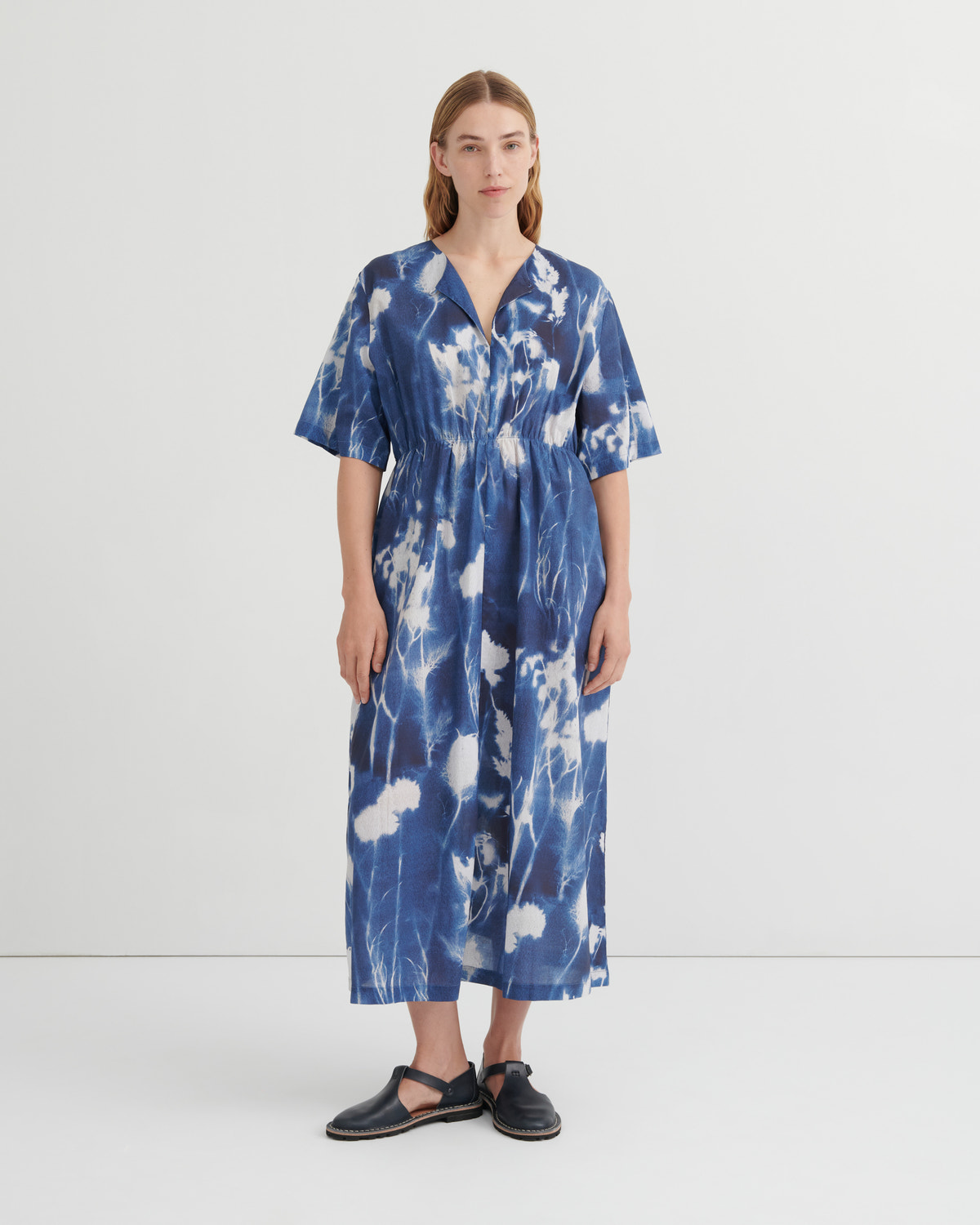 A woman stands against a plain light gray studio background wearing a loose, short-sleeve midi dress with a blue and white tie-dye or shibori-style abstract pattern. The dress has a V-neck and gathered waist.